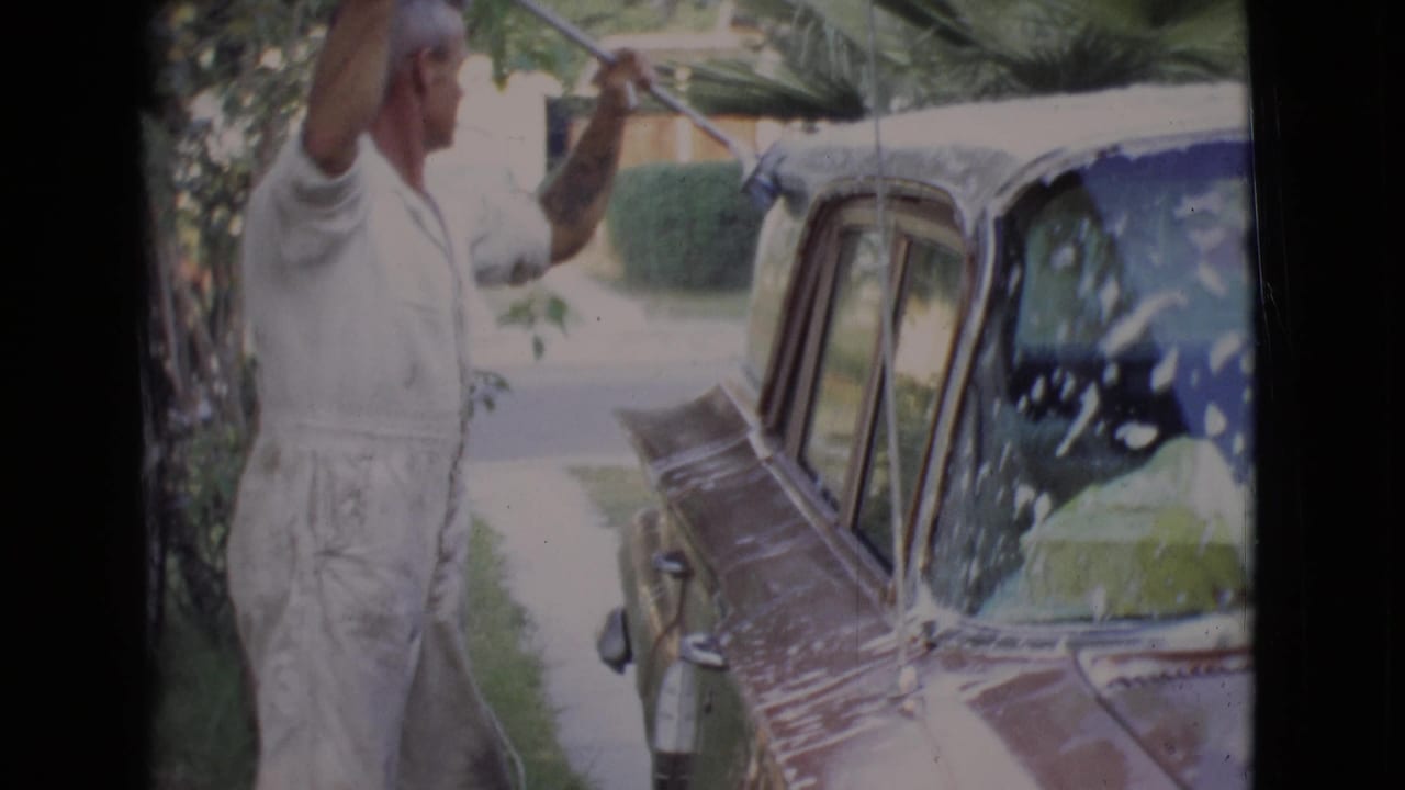 Man in white clothing washes a dark vintage car with a long-handled brush.