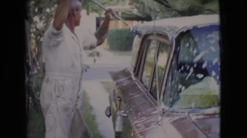 In 1968 San Diego County footage, a man in white clothing uses a long-handled brush to wash a dark vintage car near trees and a street.