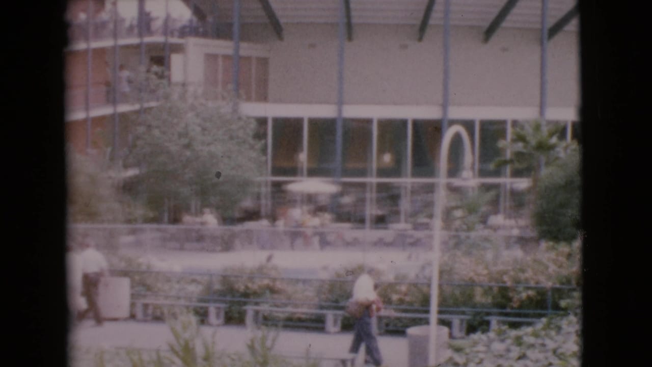 Distant pedestrians in a San Diego County public courtyard, with patio seating, landscaping, glass windows, and blue structural supports visible.