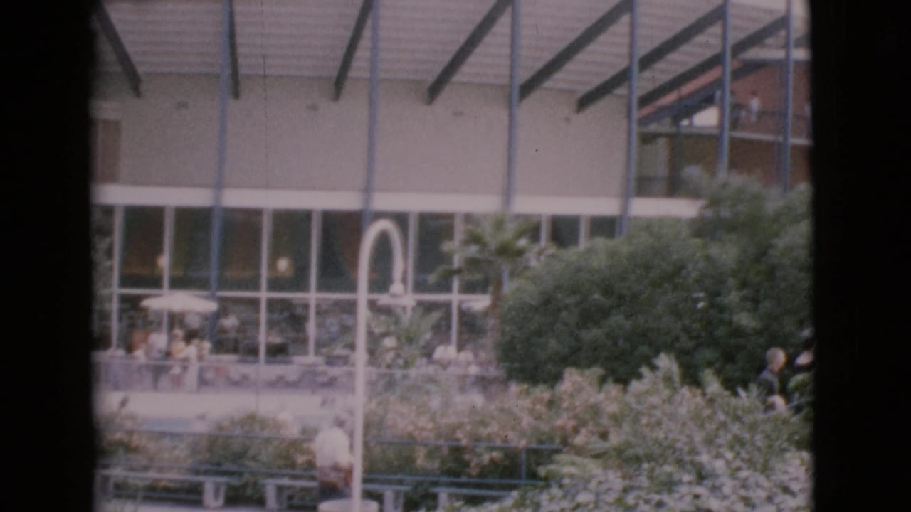 Home-movie view of a landscaped 1968 San Diego County courtyard, with distant figures, benches, glass windows, and roof trusses.