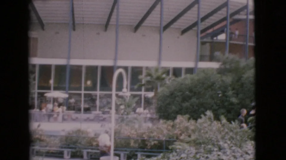Home-movie view of a landscaped 1968 San Diego County courtyard, with distant figures, benches, glass windows, and roof trusses.