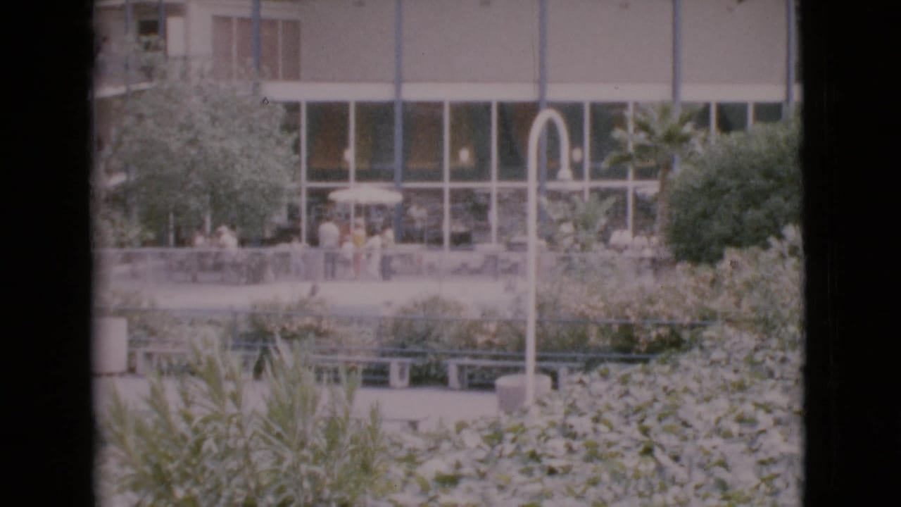 1968 San Diego County home-movie frame showing visitors near hedges and benches outside a glass-fronted mid-century building.