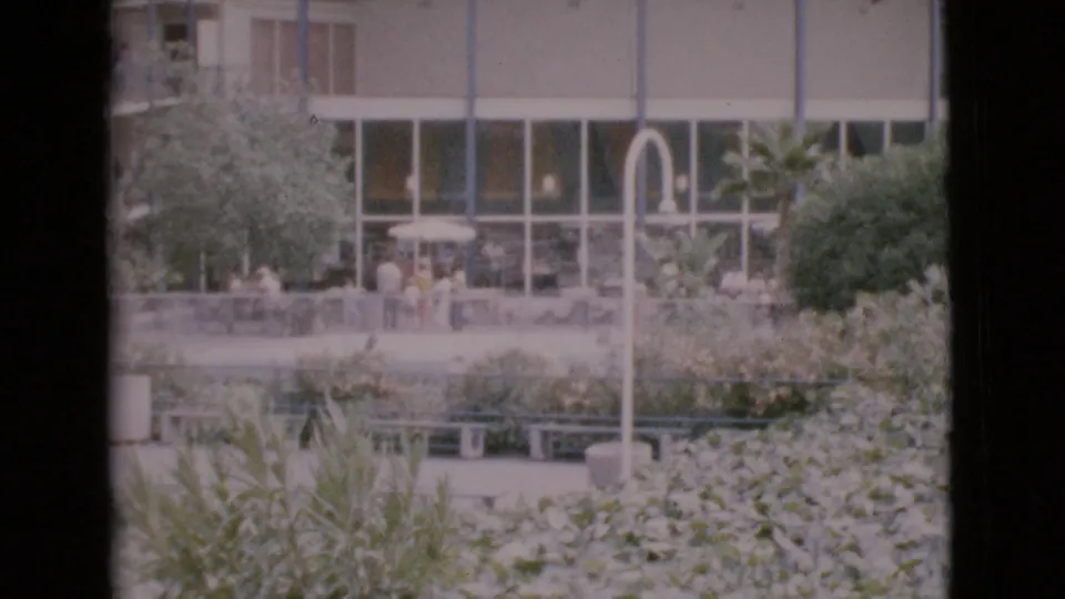1968 San Diego County home-movie frame showing visitors near hedges and benches outside a glass-fronted mid-century building.