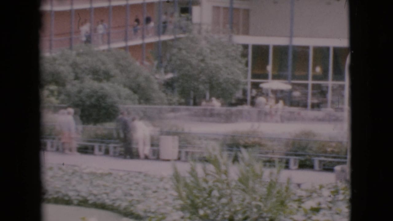 Visitors stand near benches and hedges outside a modern building with large windows.