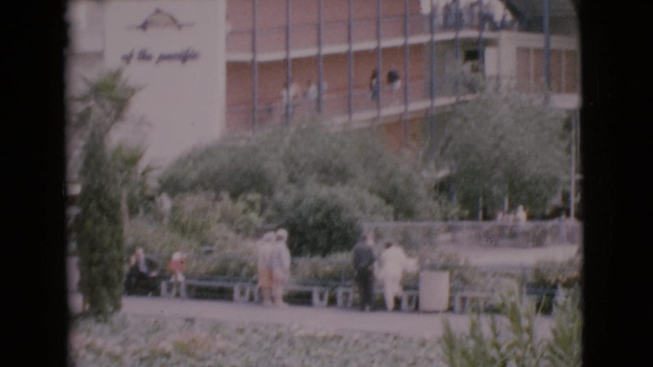 Home-movie view of visitors seated and standing in a planted courtyard beside a mid-century building in San Diego County.