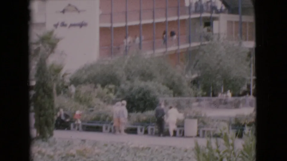 Home-movie view of visitors seated and standing in a planted courtyard beside a mid-century building in San Diego County.