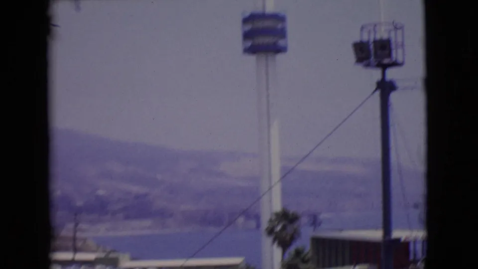 1968 San Diego County 8mm frame showing a flagpole, blue tower platform, hazy waterfront hills, and dark film borders.