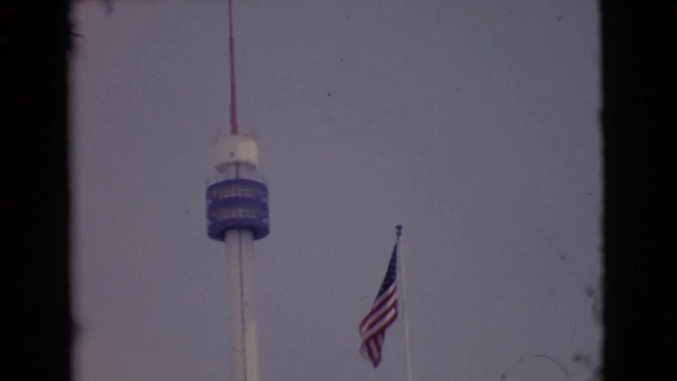 1968 San Diego County home-movie frame with a U.S. flag beside a tall tower under a muted sky.