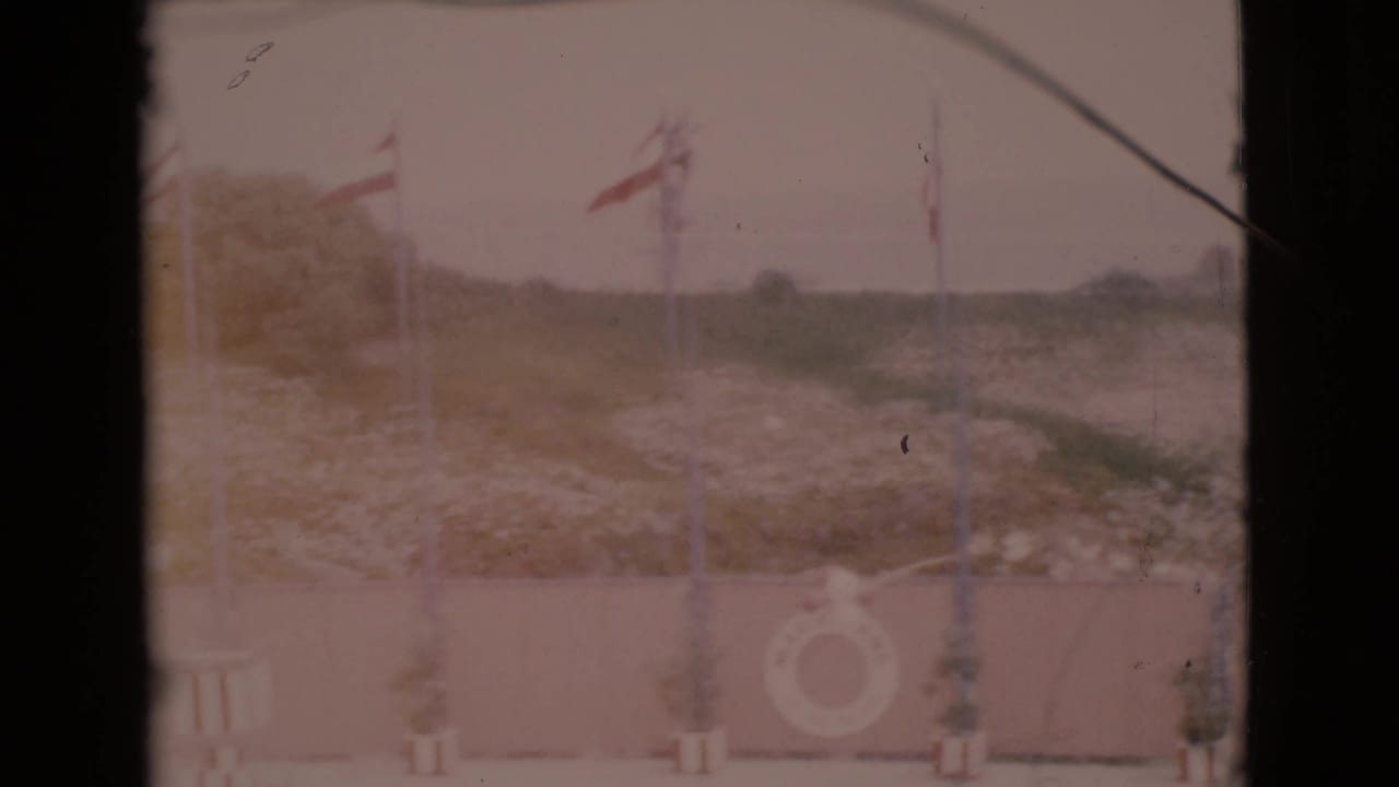 1968 San Diego County 8mm frame showing red and white flags along a low wall, with a life ring and green hillside in the background.