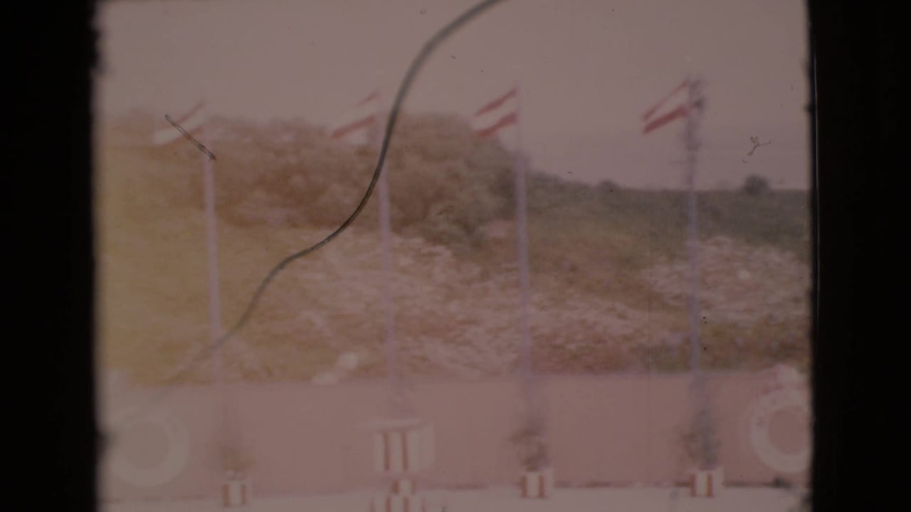 1968 San Diego County home-movie frame with red and white flags on tall poles, a pale wall, life ring, and scrubby hillside beyond.