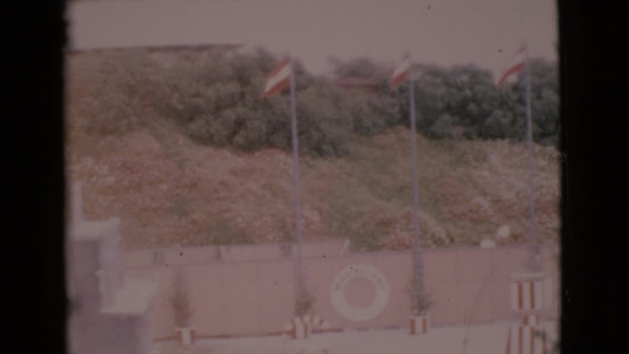 1968 San Diego County 8mm frame showing flags near a coastal wall, a visible life ring, and darker trees or shrubs behind the scene.