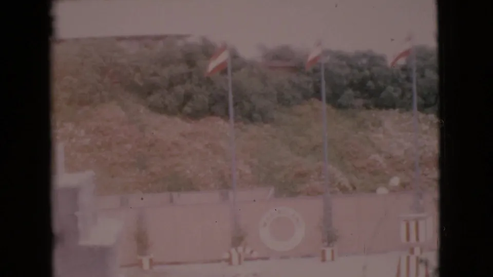 1968 San Diego County 8mm frame showing flags near a coastal wall, a visible life ring, and darker trees or shrubs behind the scene.