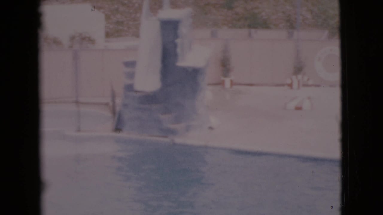 Faded 1968 home-movie frame of an outdoor pool deck in San Diego County, with steps, potted plants, a pale wall, and water along the bottom.