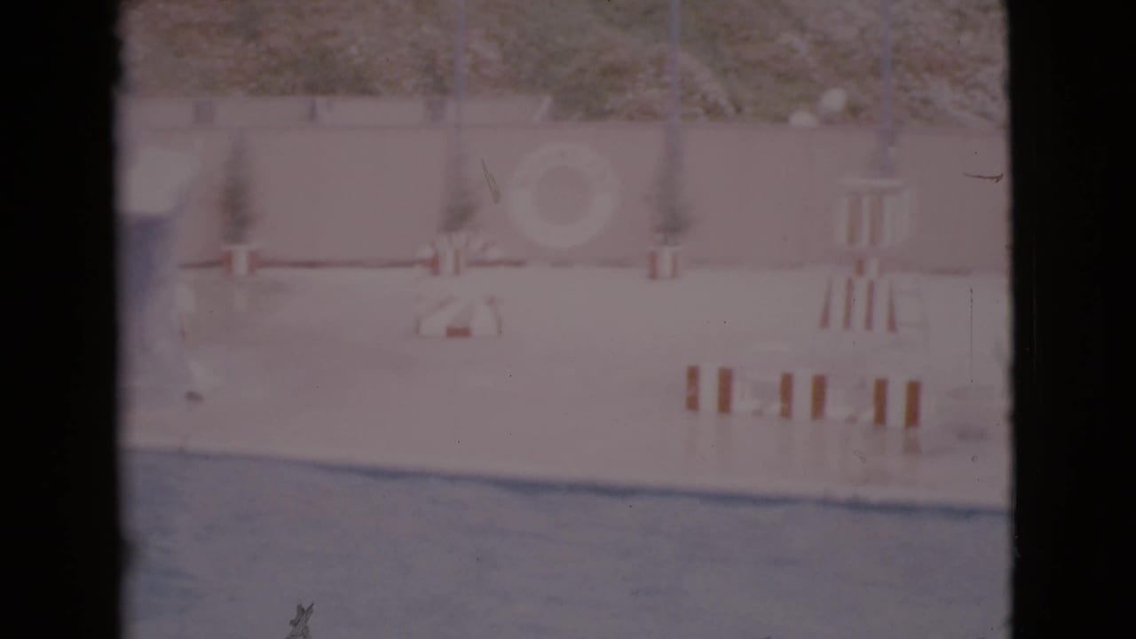 Late pan frame from 1968 San Diego County footage showing the pool wall, safety ring, small chair or stand, deck objects, hillside, and blue water.