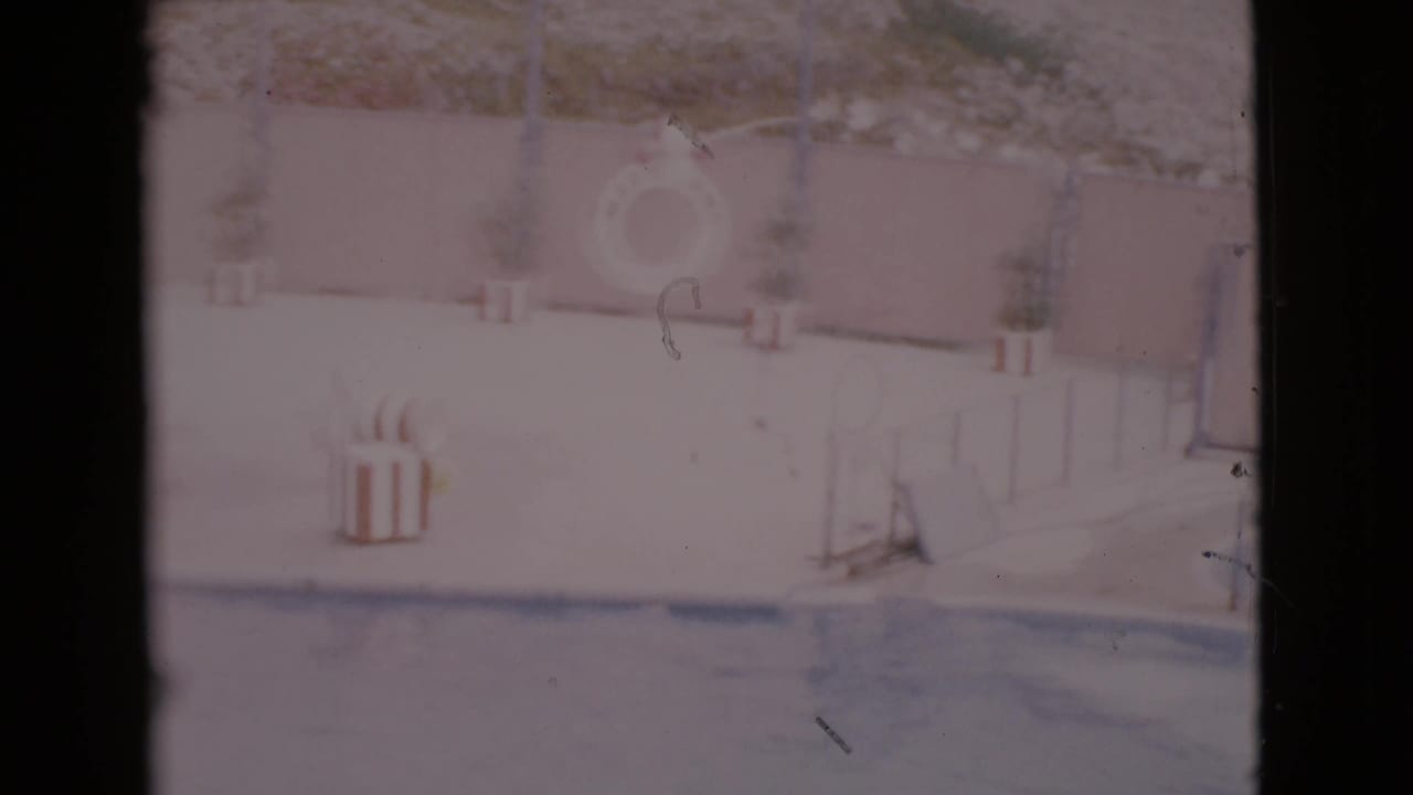 Faded 1968 San Diego County home-movie frame of a poolside area with a life ring, deck furniture, and low railing.
