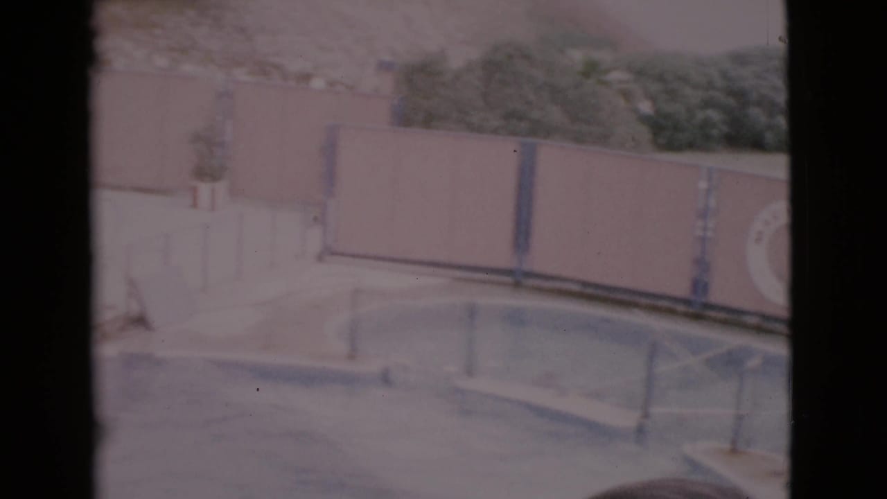 1968 San Diego County 8mm view of a curved pool edge with concrete deck, wall panels, railings, and plantings beyond.
