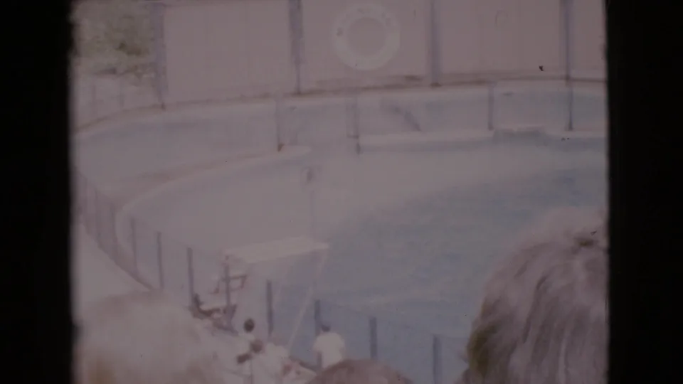 Faded 8mm frame of the Marineland show pool with a platform, railings, audience heads, and activity near the performance area.