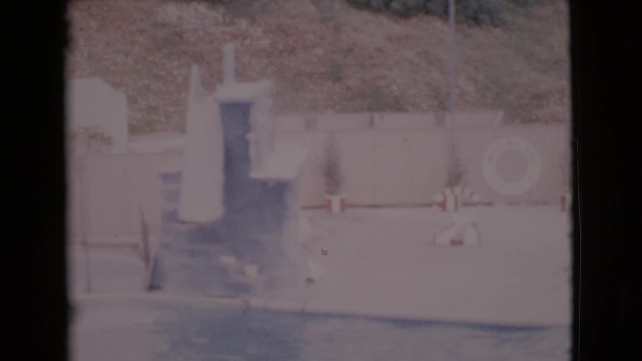 Empty pool area with a diving board, wall life ring, fence, and hillside beyond.