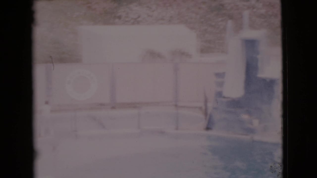 1968 San Diego County home-movie view of a quiet pool area, showing a life ring, diving board, low fencing, and pale poolside walls.