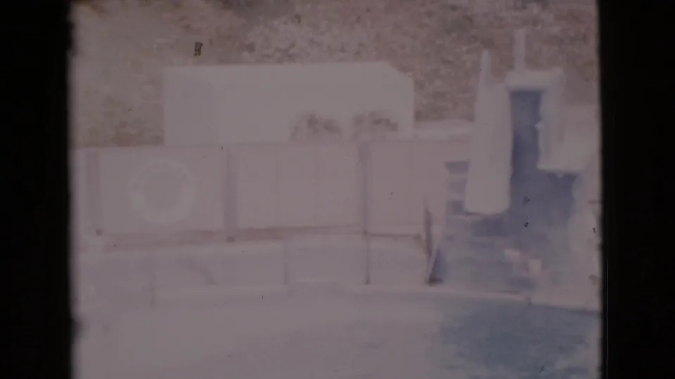 Faded 8mm frame from 1968 San Diego County showing an empty pool area with a mounted life ring, diving board, low wall, and hillside.