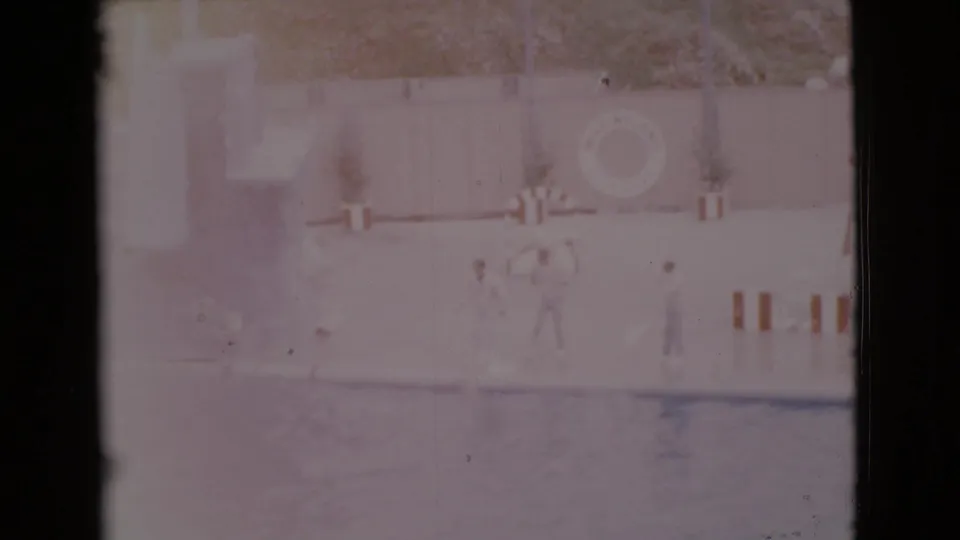 1968 San Diego County poolside home-movie frame with a dog on a surfboard, distant people, striped furniture, and a circular life preserver.