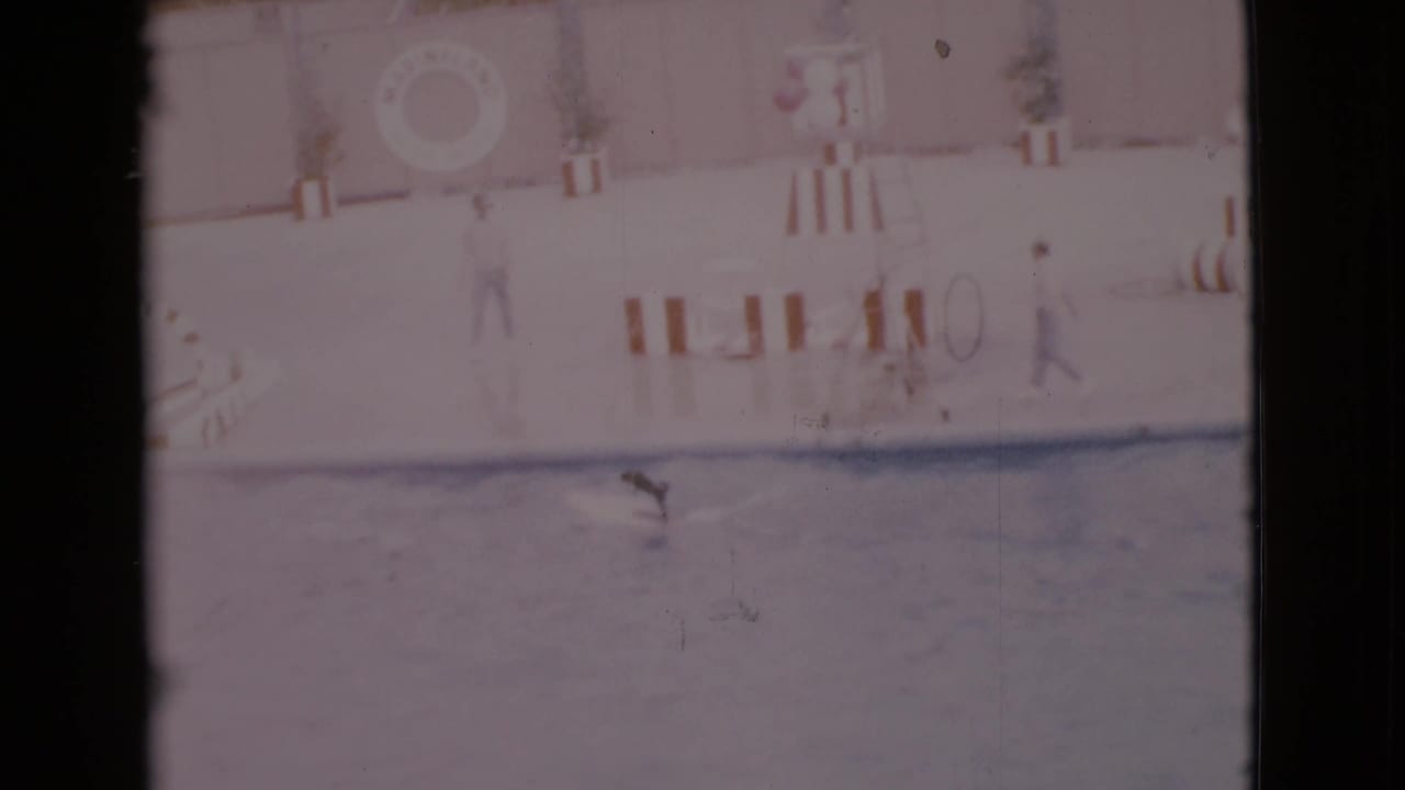 1968 San Diego County home-movie frame showing a pale indoor pool, a small swimmer, adults on deck, striped barriers, and a life ring.