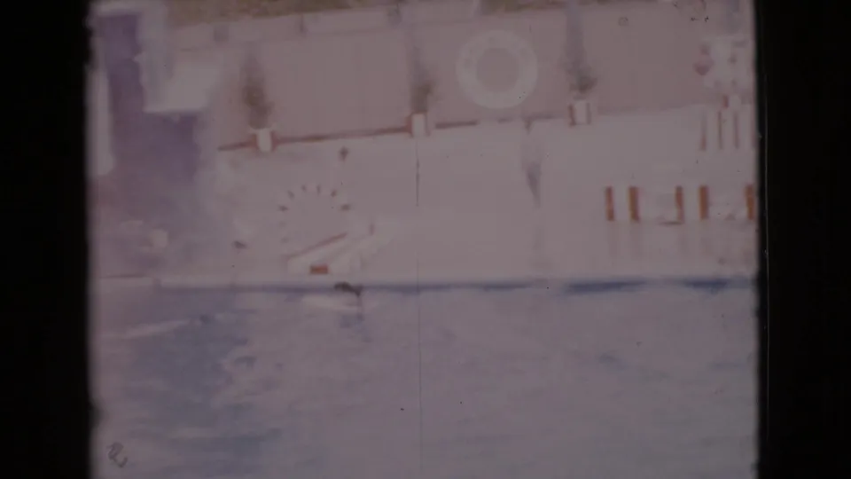 San Diego County 1968 8mm frame showing a small swimmer in an indoor pool with adults, deck markings, potted plants, and film scratches.