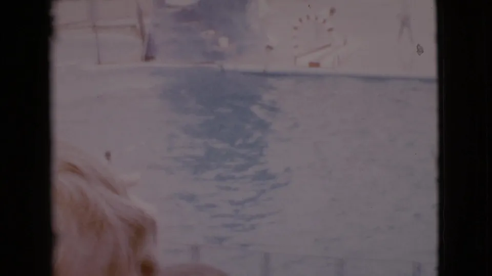 Soft, overexposed 1968 home-movie frame with blond hair at lower left, rippled water, and pale background structures.