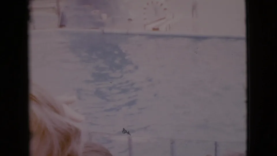 Faded 8mm San Diego County frame with rippled water, a pale pool or lagoon edge, and blond hair low in the foreground.