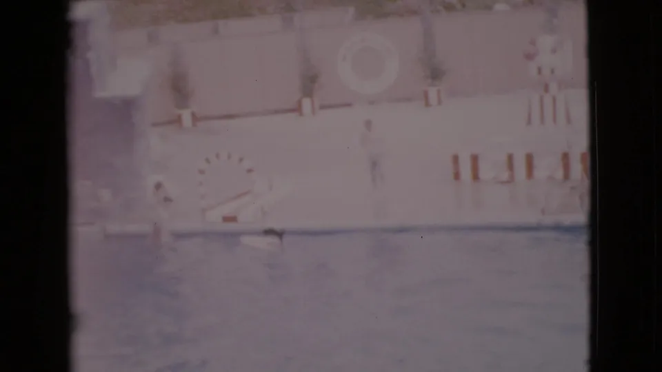 1968 San Diego 8mm frame showing a public aquarium pool, striped hoop, wall life preserver, and trainer during a dolphin show.