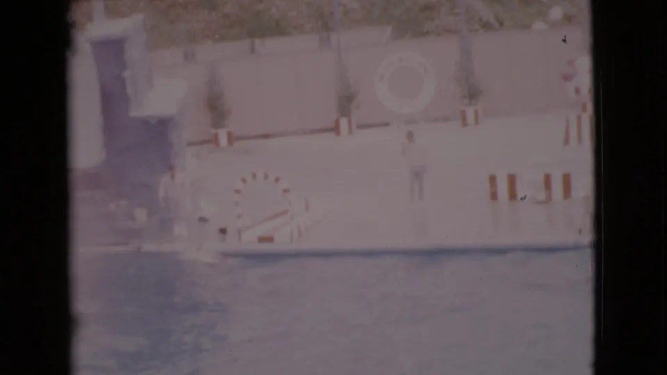 1968 San Diego home-movie frame of a dolphin show area with trainer, hoop, pool, wall life preserver, and plants.