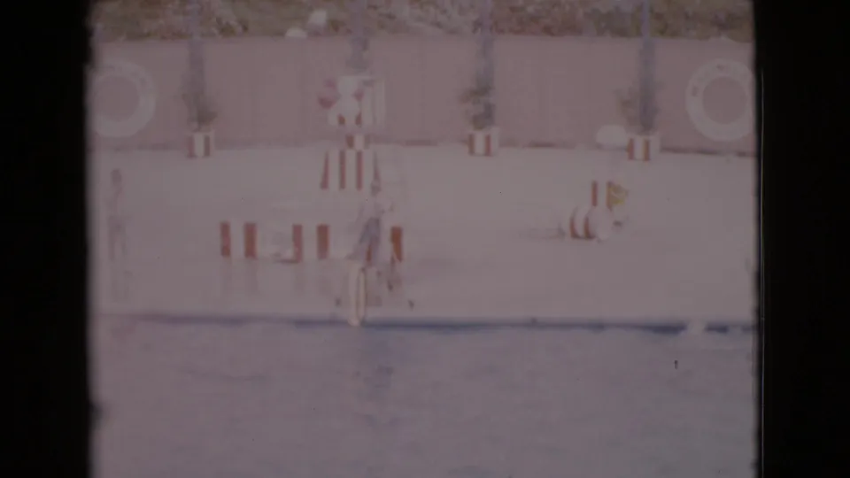 1968 San Diego County 8mm frame showing a dolphin performance pool with a trainer near hoop props and deck barriers.