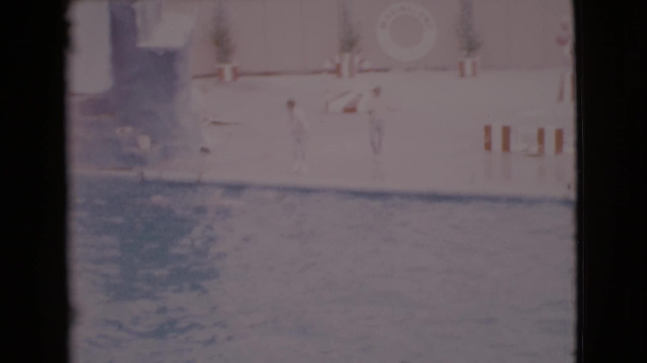 1968 San Diego County home-movie frame showing a marine show pool, a dolphin splash, and poolside barriers in faded 8mm color.
