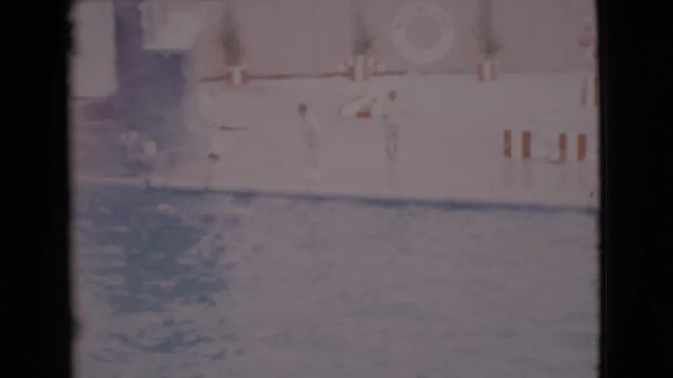 1968 San Diego County home-movie frame showing a marine show pool, a dolphin splash, and poolside barriers in faded 8mm color.