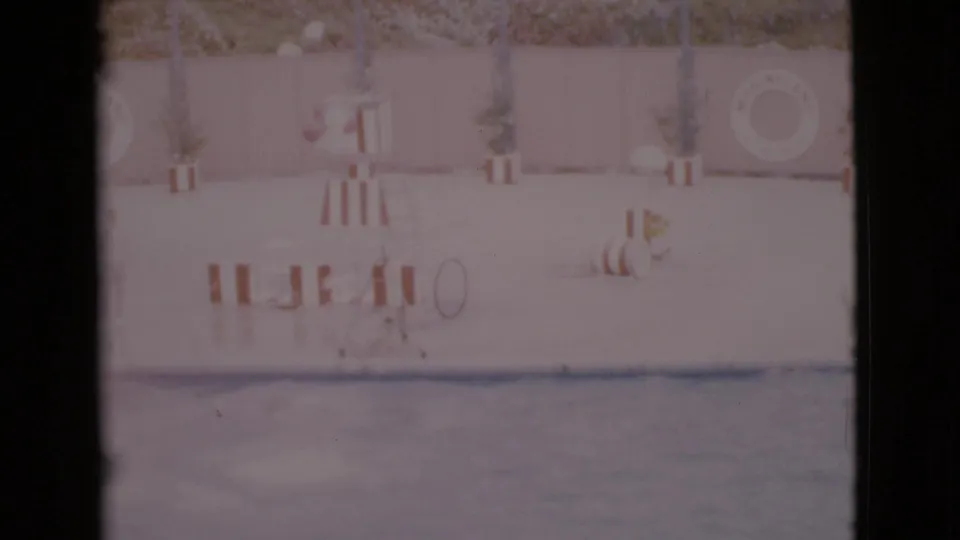 1968 San Diego County home-movie frame showing a sunny pool area with a striped stand, wall-mounted life ring, potted plants, and water in front.