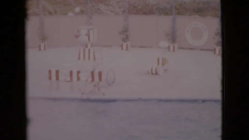 Soft 1968 San Diego County home-movie frame showing pool water, a wall-mounted life ring, striped stand, potted plants, and faint people nearby.