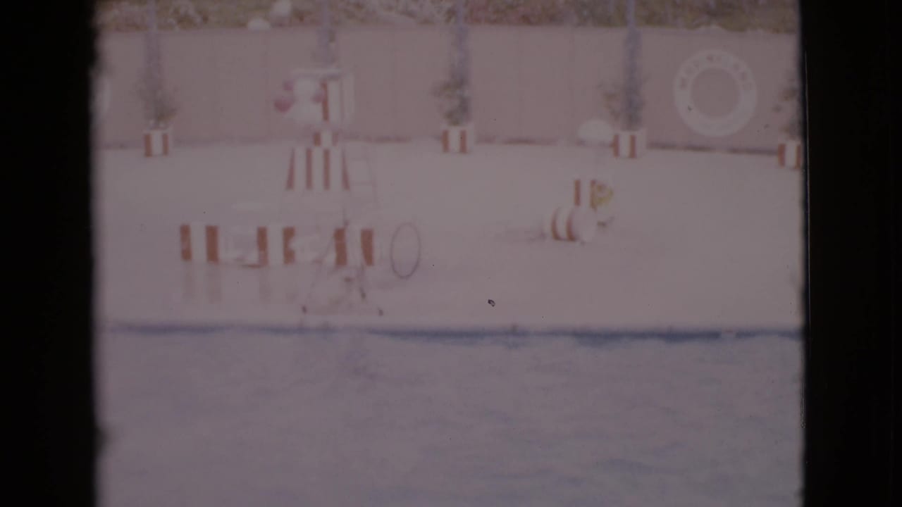 Empty poolside view with striped furniture, lifeguard stand, potted plants, and a life ring on the wall.