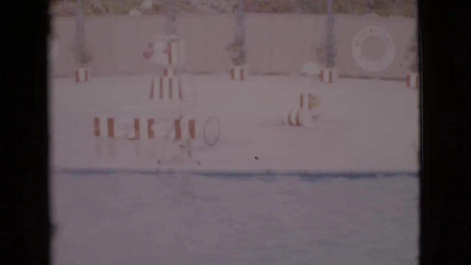 1968 San Diego County 8mm frame showing an empty swimming pool area with striped furniture, a stand, plants, and a wall-mounted life ring.