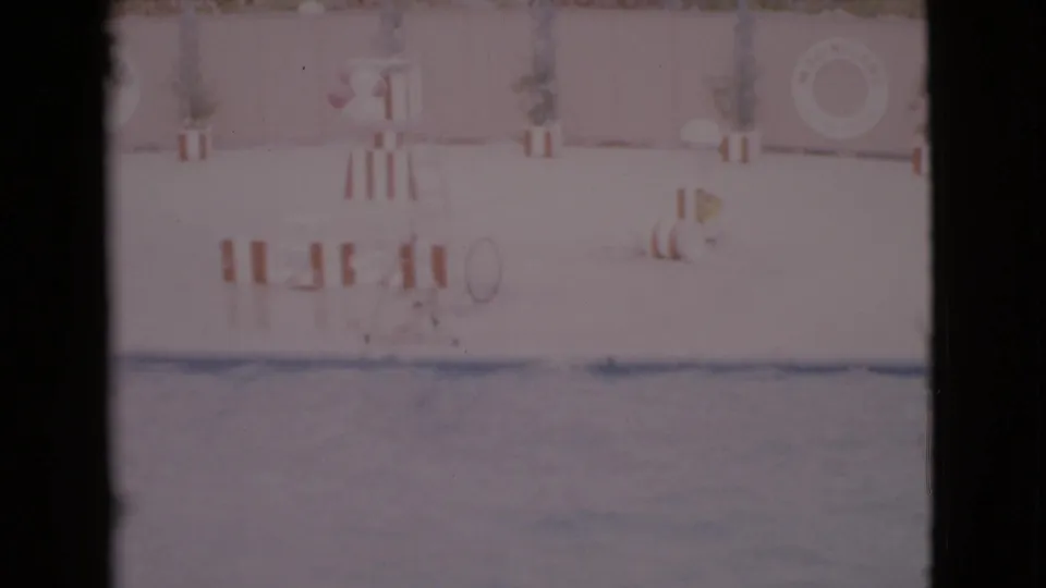 Faded 8mm view of the San Diego County pool area, with striped seating, potted plants, a stand, and a life ring against the wall.
