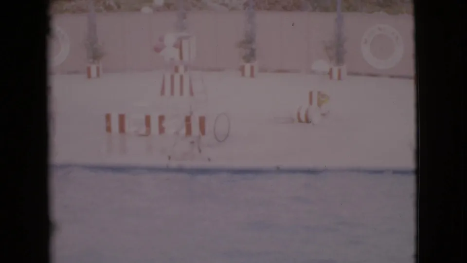 1968 home-movie frame of a quiet San Diego County pool, with water in the foreground and striped furniture, plants, and a life ring behind it.