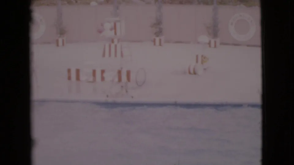 Faded 8mm frame from San Diego County showing an empty poolside setting with striped furnishings, plants, a stand, and a wall life ring.