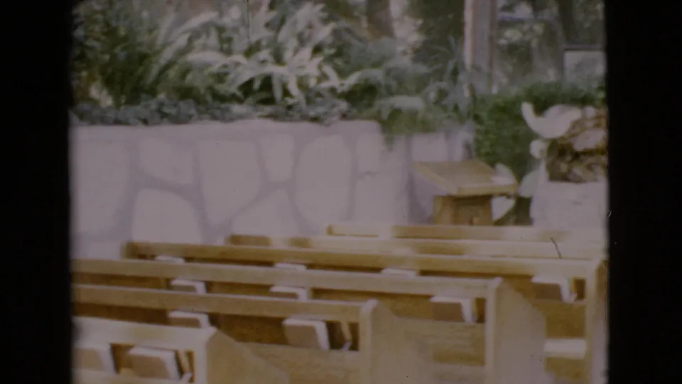 1968 San Diego County frame showing pew rows, indoor greenery, a pale stone wall, and part of a religious interior.