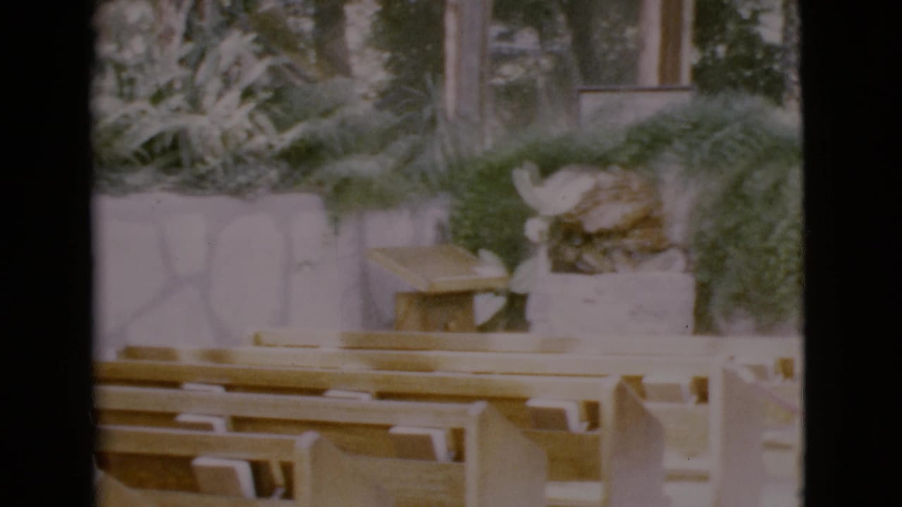 1968 San Diego County 8mm frame showing empty wooden pews, greenery, and a cross near a white Catholic church altar area.