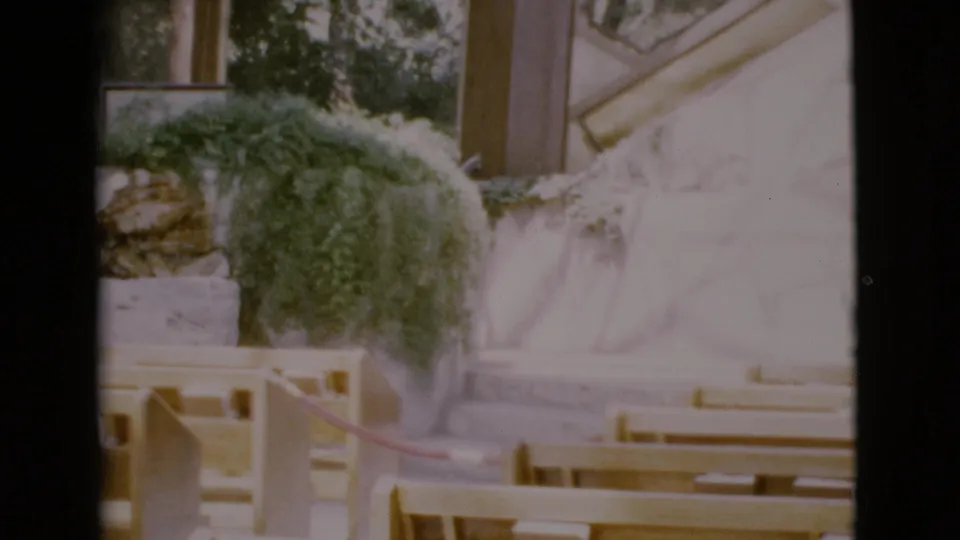 Vintage 8mm frame of an empty Catholic church interior in San Diego County, with pews, plants, stonework, and a pale altar wall.