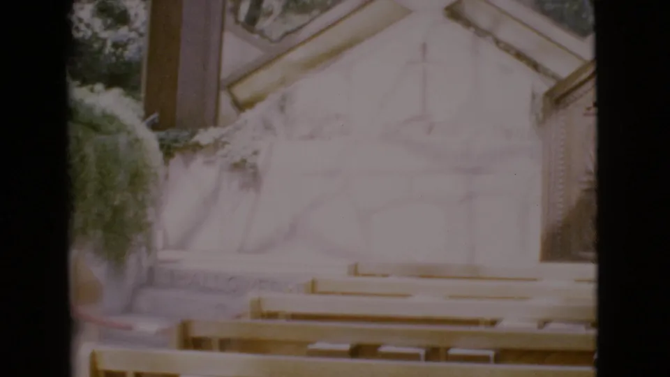 End frame of the 1968 San Diego County church interior, showing empty pews, a pale altar wall, greenery, and soft 8mm exposure.