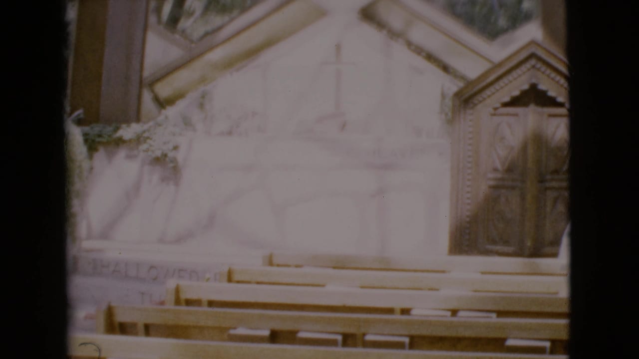 Church interior with pews facing a pale altar wall, cross, and wooden doors.