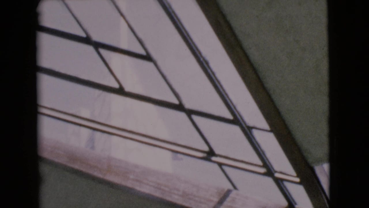1968 San Diego County 8mm frame showing angled glass panes, dark mullions, a wood beam, and a pale textured wall in daylight.