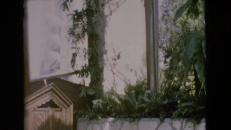 1968 home-movie frame of the lower church area with plants, a stone wall, and a small wooden pulpit or lectern.