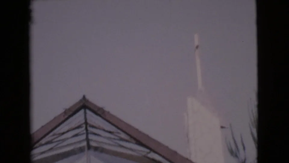 1968 San Diego County frame showing the church roof peak, steeple, pale sky, and edge foliage.