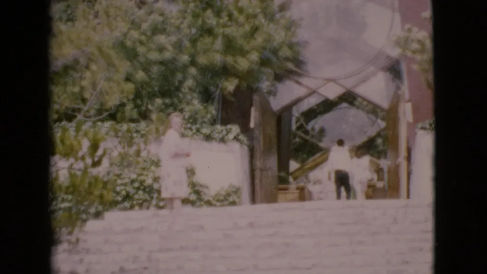 1968 San Diego County home-movie frame showing a wide staircase, trees, and two adults near a geometric building entrance.